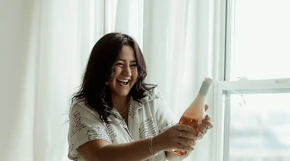 Claire Smith in front of a window joyfully opening a bottle of bubbly wine.